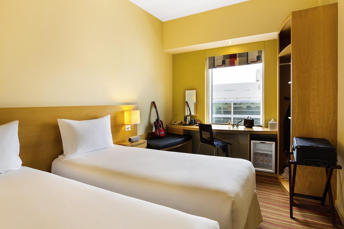 A hotel room with two beds, a desk, a chair, a wardrobe, and a window with a view of buildings.