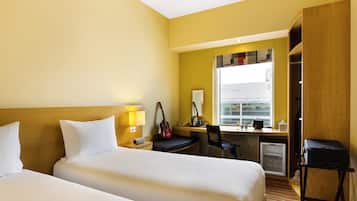 A hotel room with two beds, a desk, a chair, a wardrobe, and a window with a view of buildings.