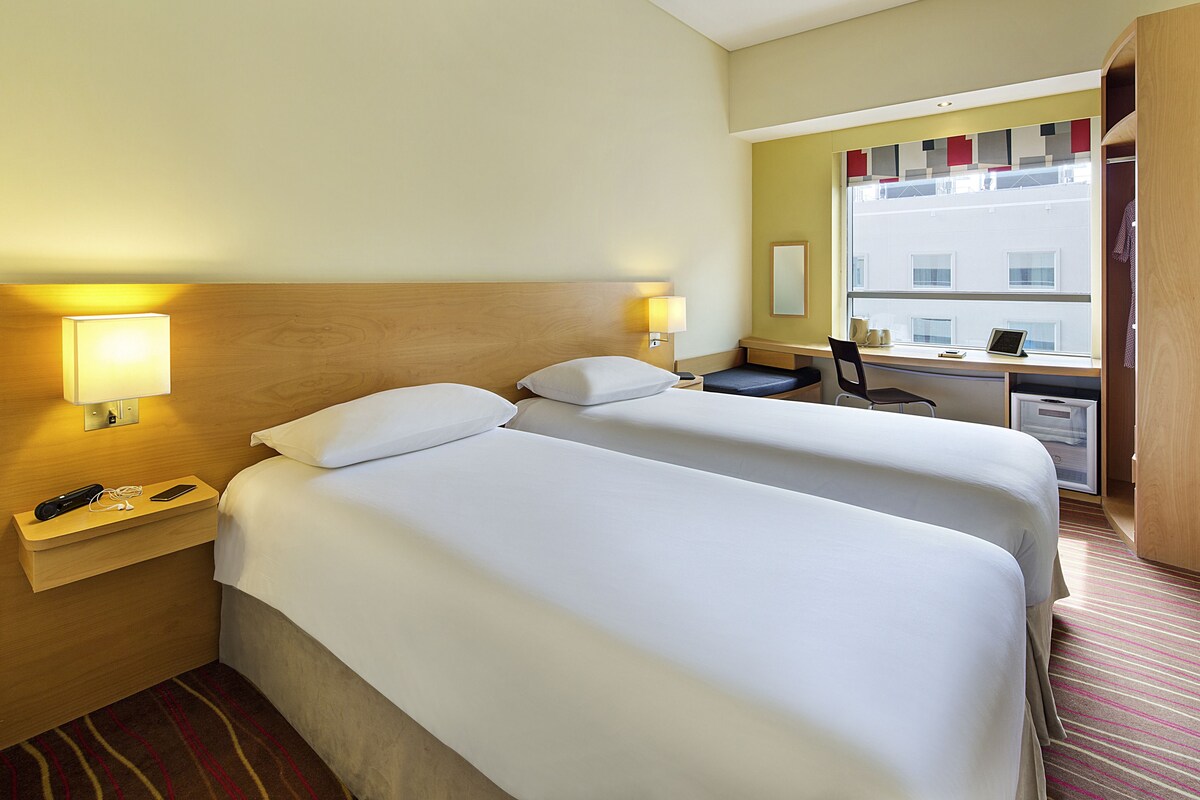 A hotel room with two beds, a desk, a chair, and a window with a view of buildings.
