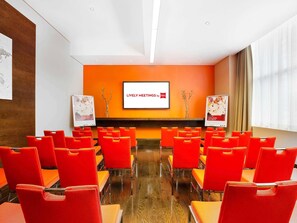 A conference room with orange chairs, a large screen displaying 'Lively Meetings by ibis,' and two posters on stands. - ibis Dubai Al Rigga (Dubai)