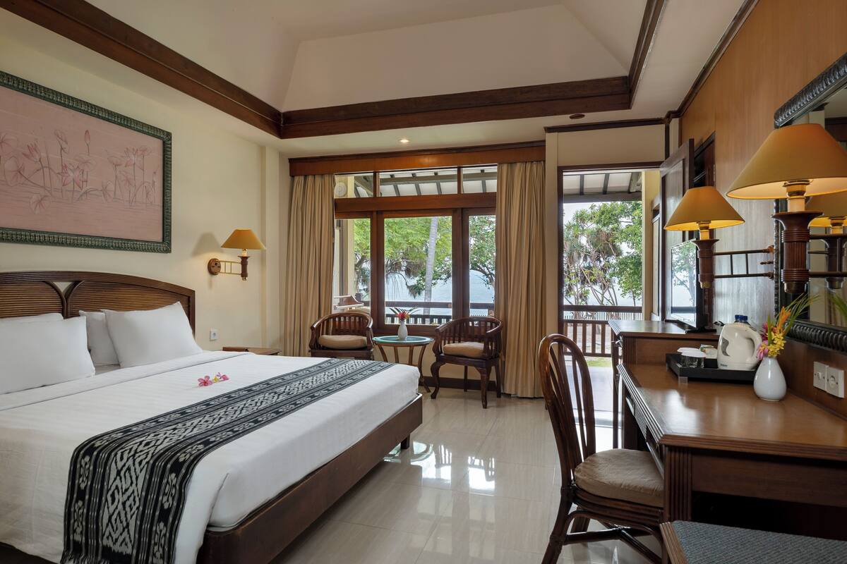 A hotel room with a large bed, a desk, two chairs, a small table, and a balcony with curtains.