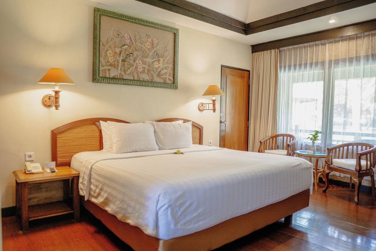 A neatly made bed with white linens, a wooden headboard, and two bedside lamps. There is a small table with a chair and a painting on the wall above the bed.