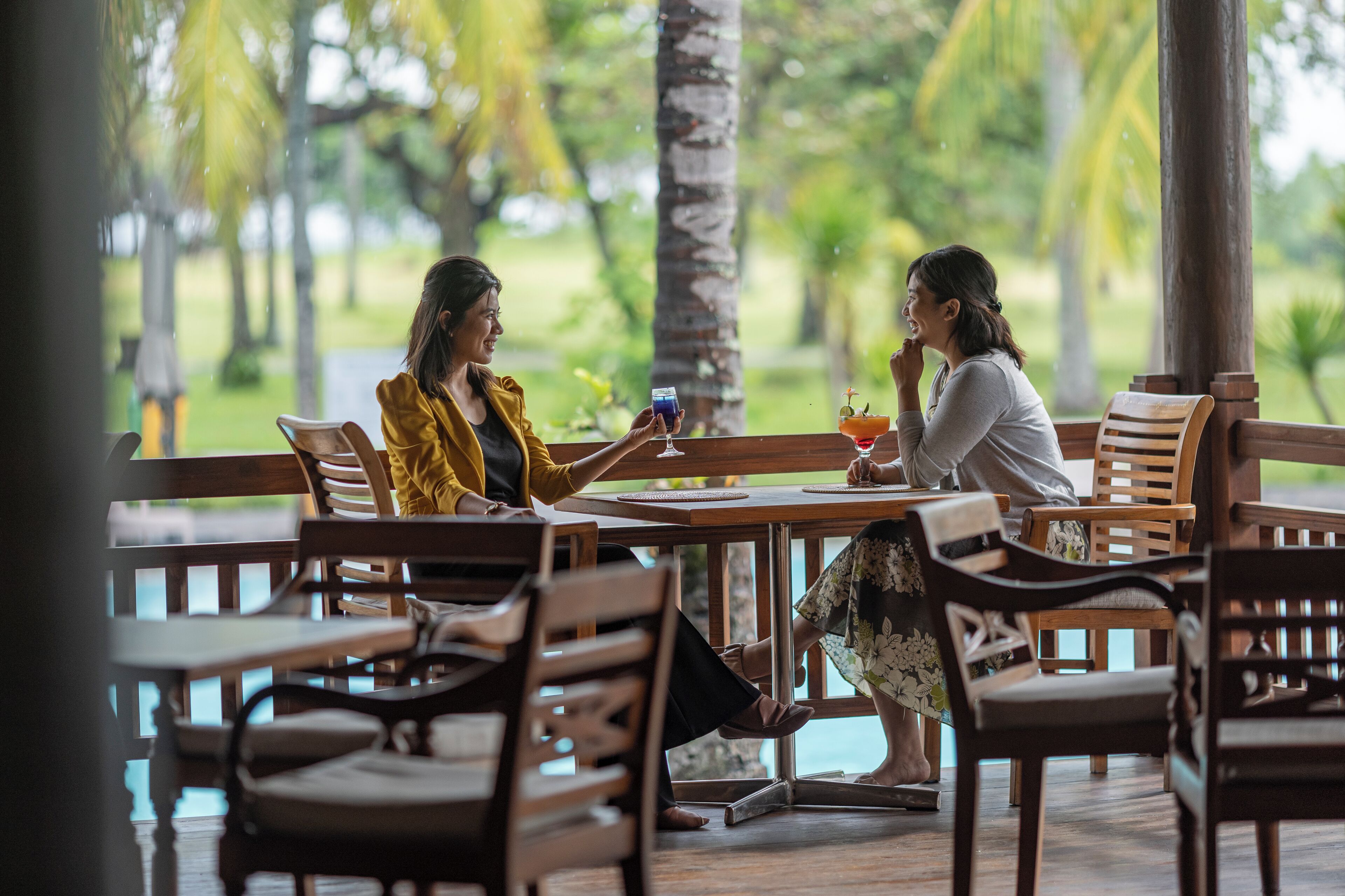 Two people seated at an outdoor wooden table with a view of a lush garden and a pool.