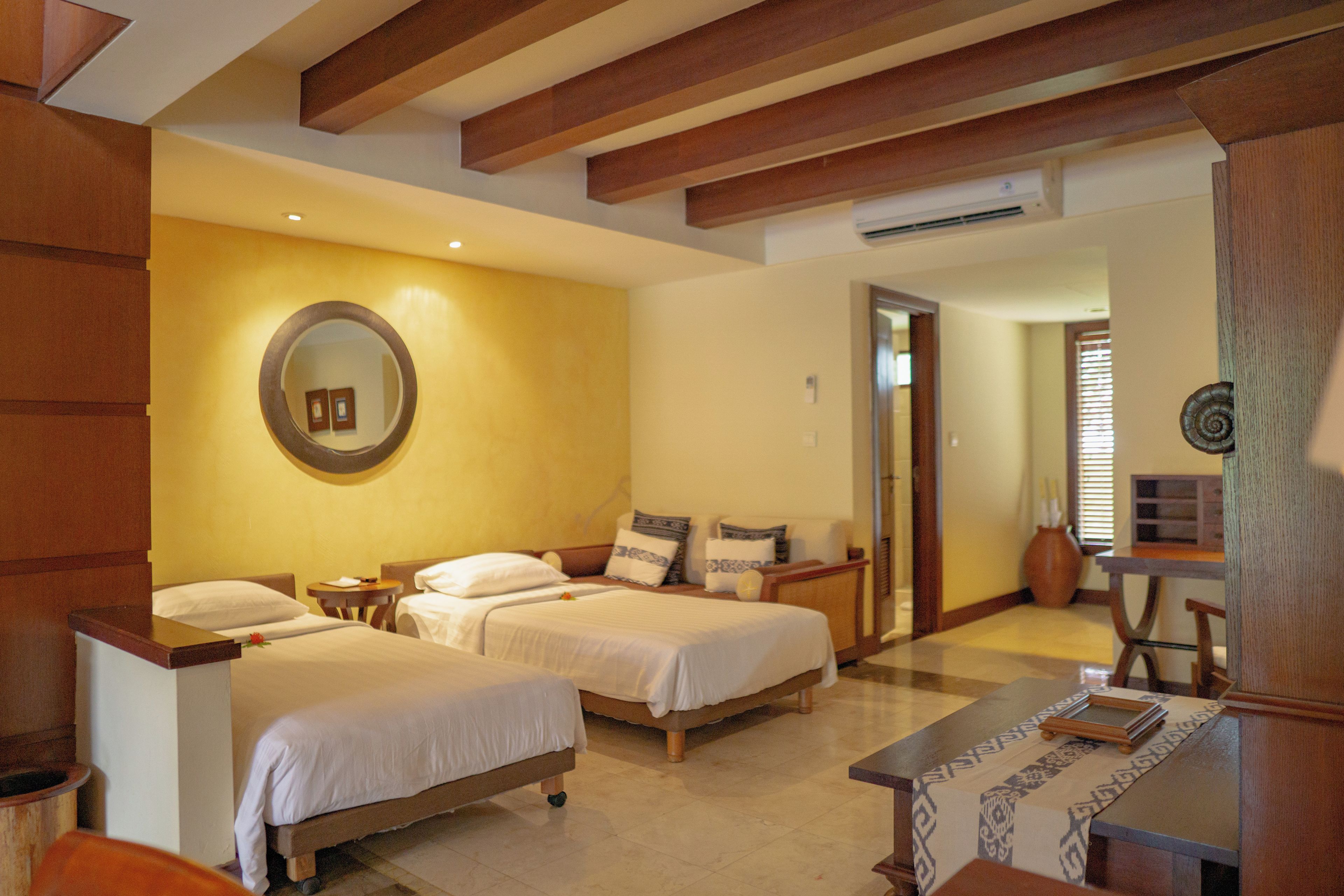 A hotel room with two beds, a wooden headboard, a round mirror, a small table, and a chair.