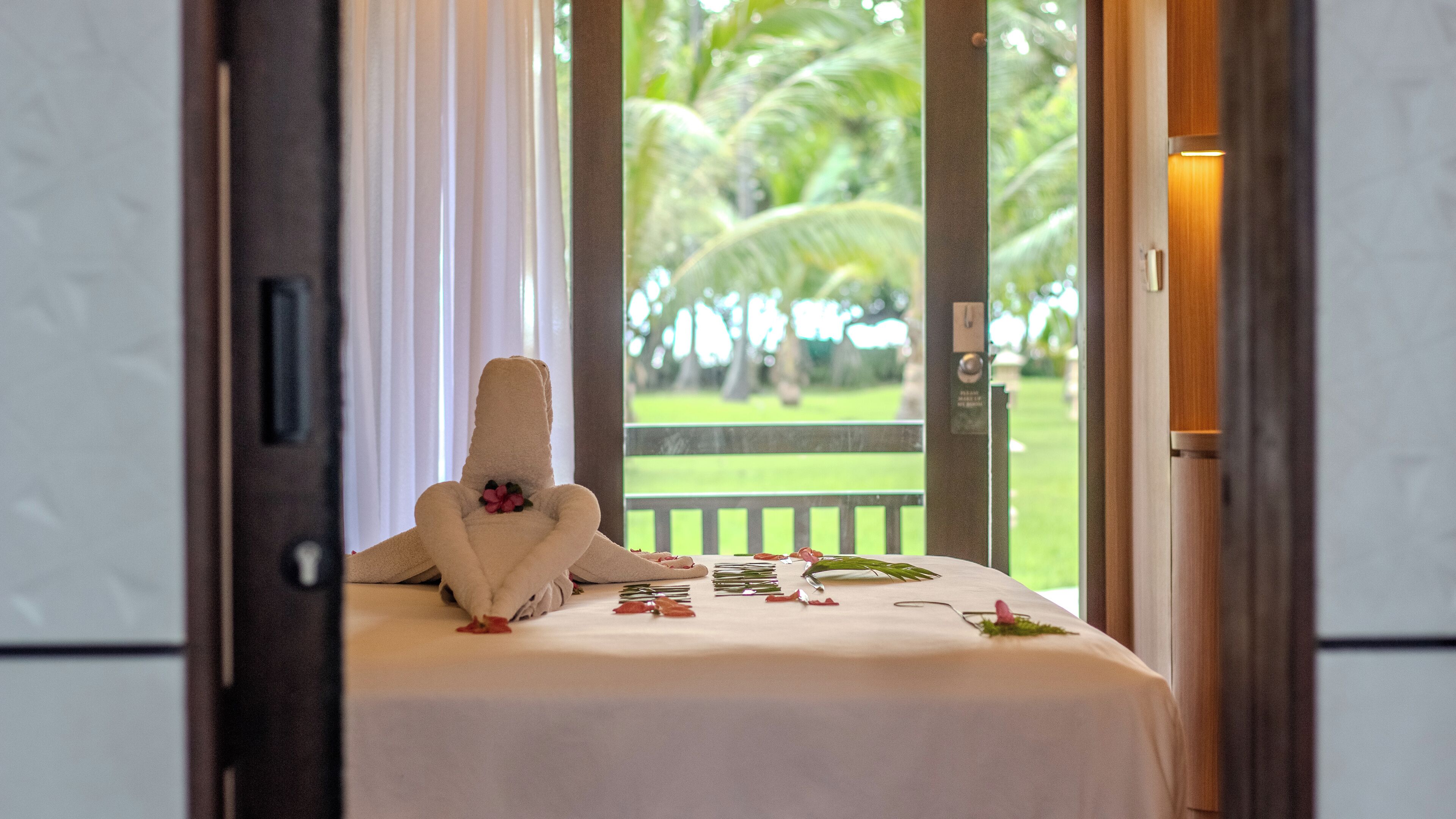 A hotel room with a bed, a towel, and a view of a green landscape with palm trees.