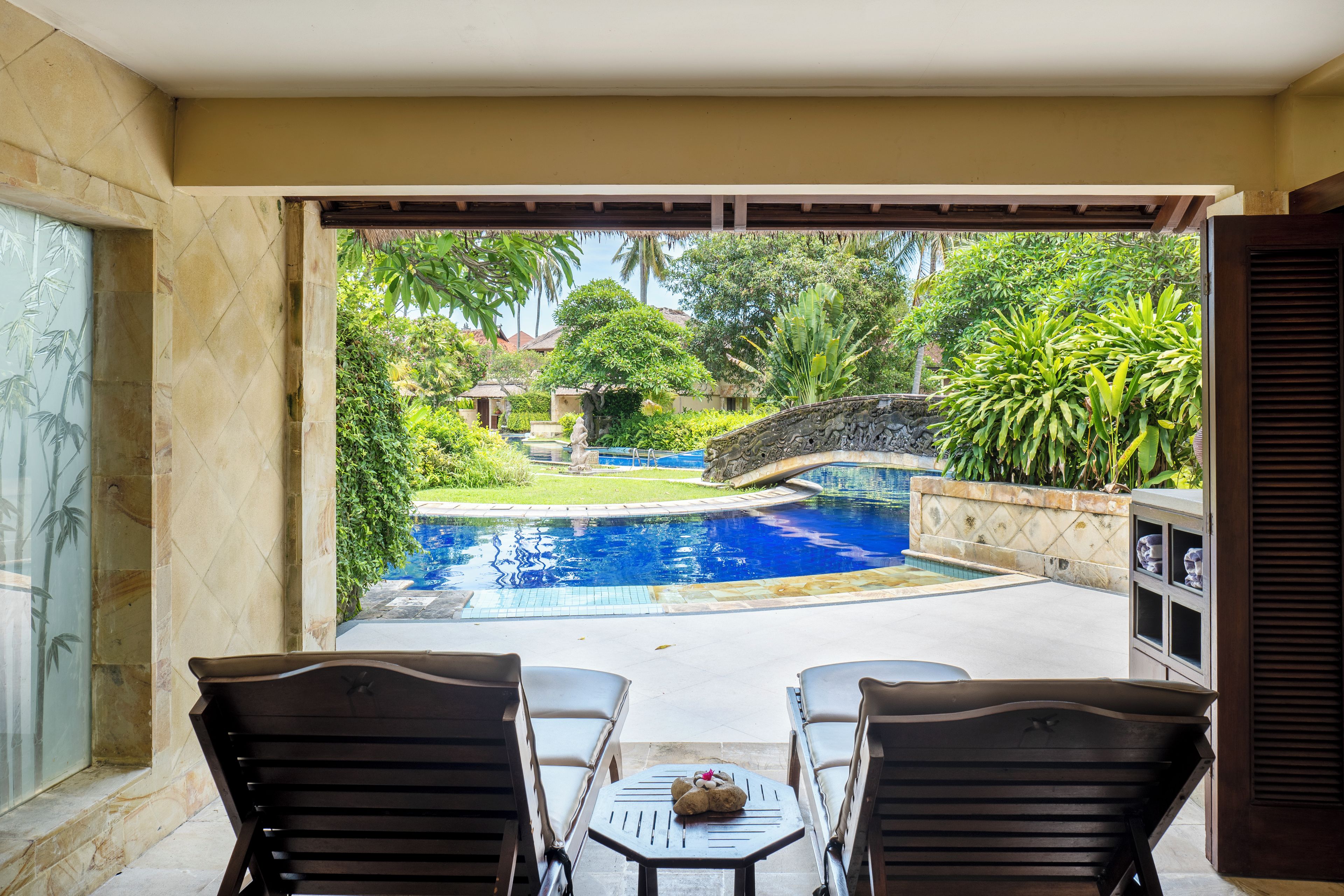 A pool area with a stone bridge, surrounded by lush greenery and a thatched roof structure.