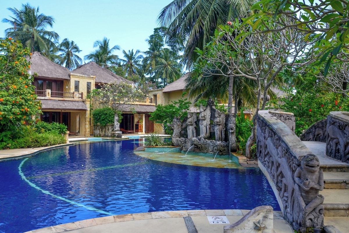 A swimming pool with a stone bridge, surrounded by tropical vegetation and traditional architecture.