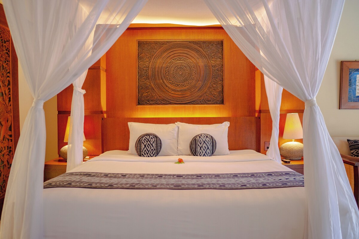 A hotel room with a large bed, white bedding, a decorative headboard, and a nightstand with a lamp.