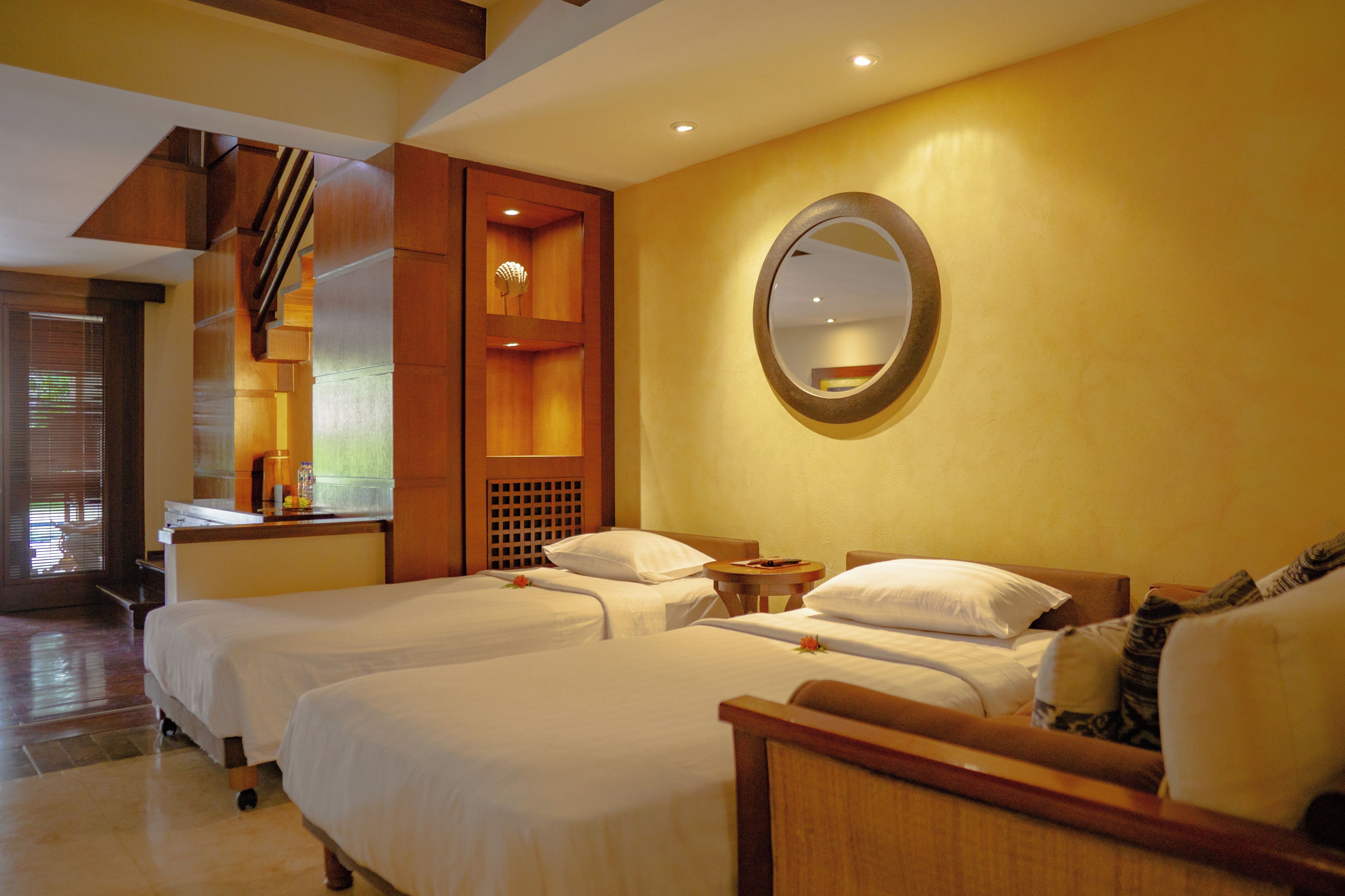 A hotel room with two beds, a wooden headboard, a round mirror, and a small table with a lamp.