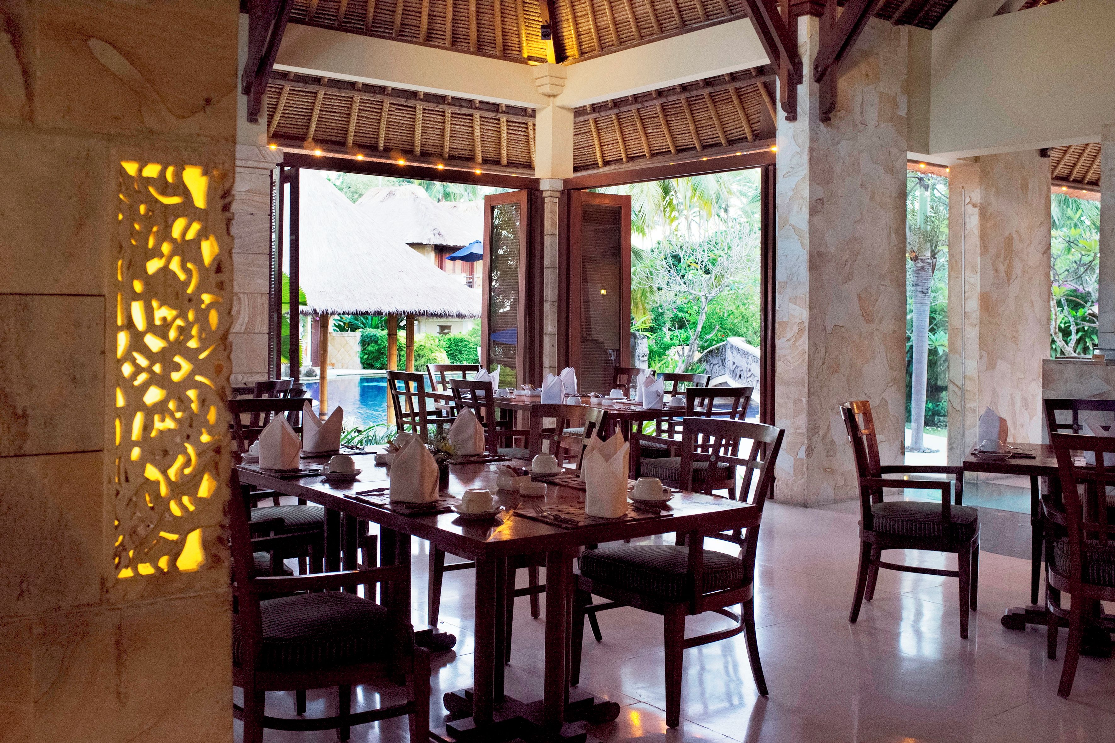 A restaurant with multiple tables set for dining, a pool visible through the windows, and a tropical garden view.