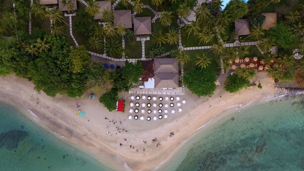 Vista aérea de un complejo turístico en la playa con un edificio central rodeado de palmeras, una playa de arena y aguas turquesas cristalinas.