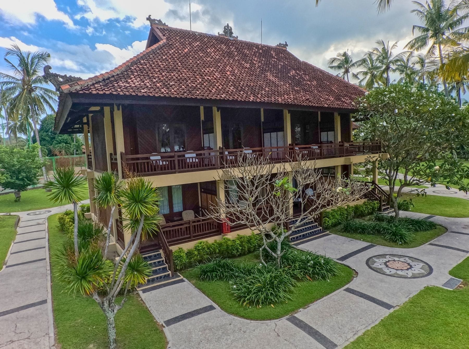 A two-story building with a tiled roof, a balcony with seating, and a well-maintained garden with palm trees and shrubs.