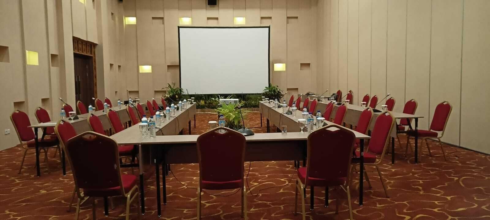 A conference room with a long table, red chairs, and a projector screen.