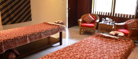 A room with two single beds, a wicker chair with a red cushion, a small table with a glass top, and a window with vertical blinds.