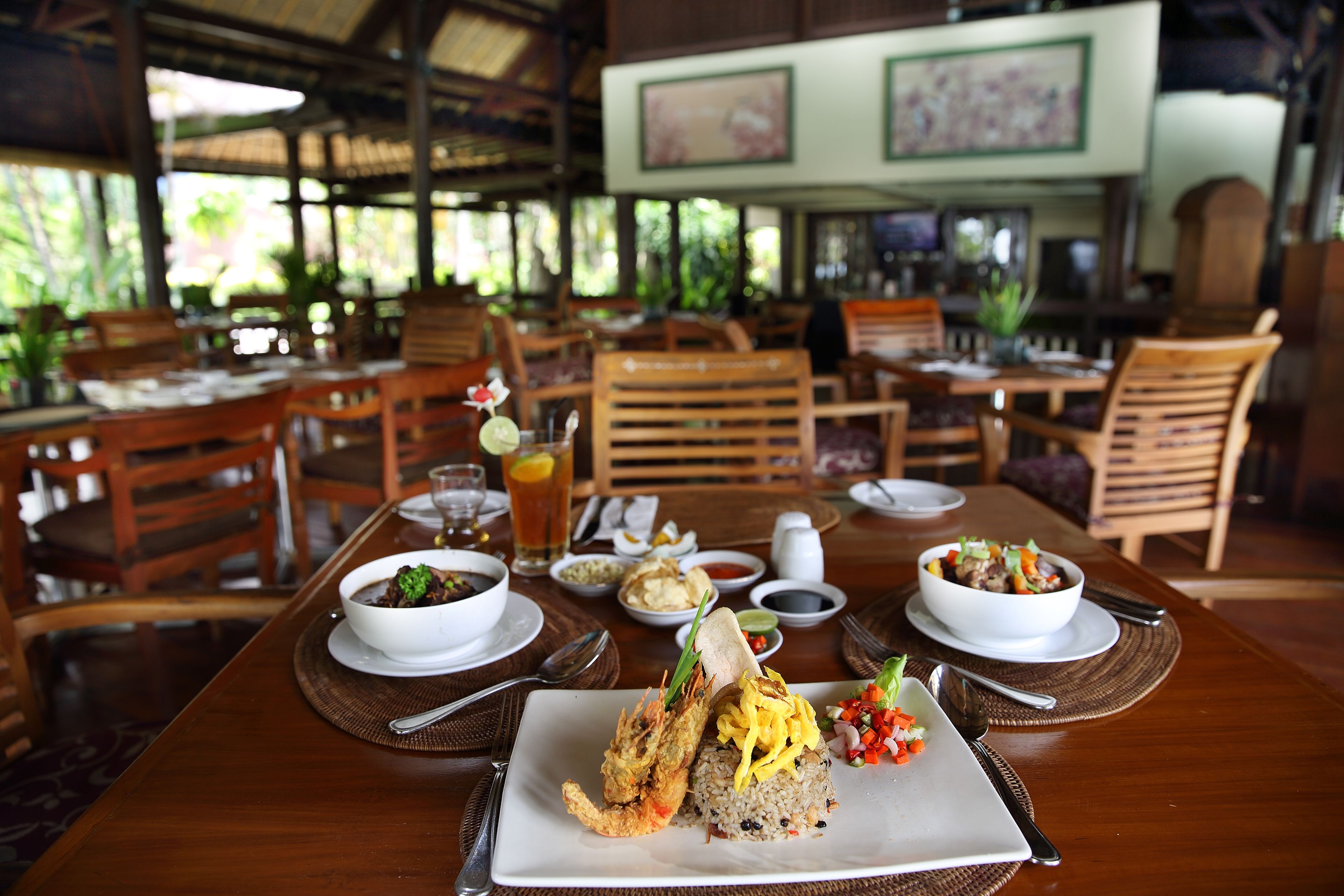 A restaurant with wooden tables and chairs, a variety of dishes including fried shrimp and rice, and a tropical setting with plants and a thatched roof.