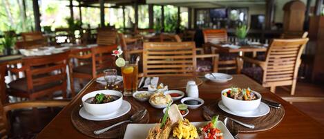 A restaurant with wooden tables and chairs, a variety of dishes including fried shrimp and rice, and a tropical setting with plants and a thatched roof.