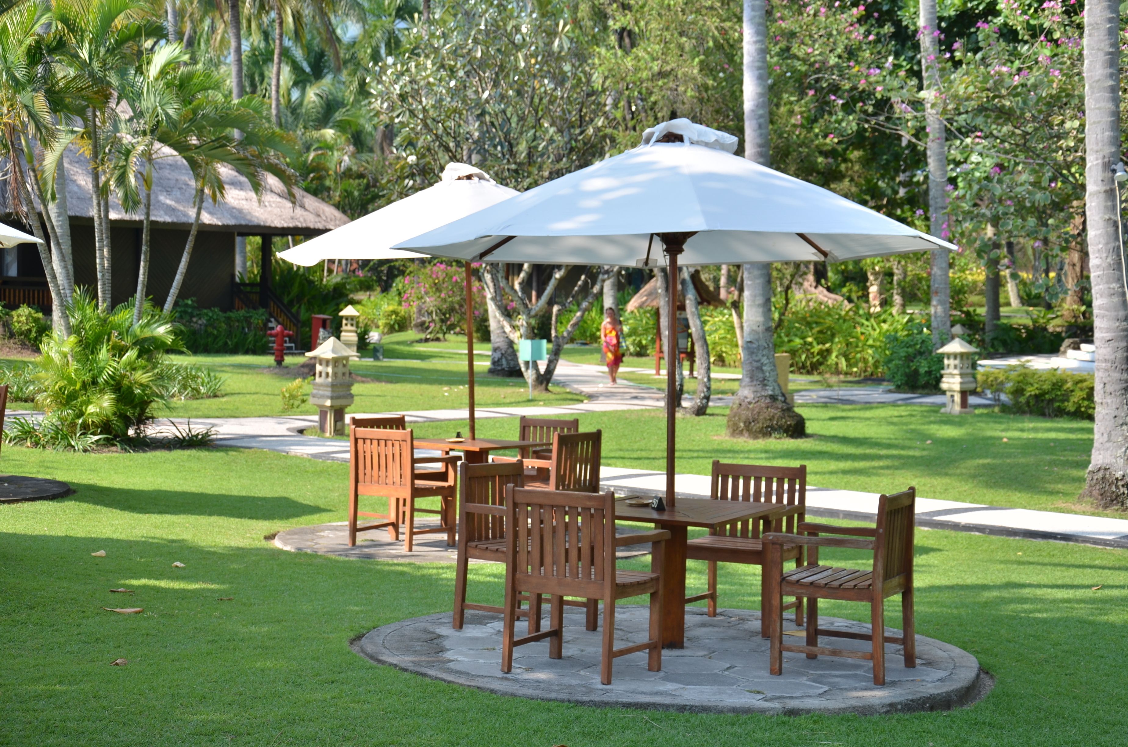 An outdoor seating area with wooden tables and chairs, surrounded by lush greenery and palm trees.