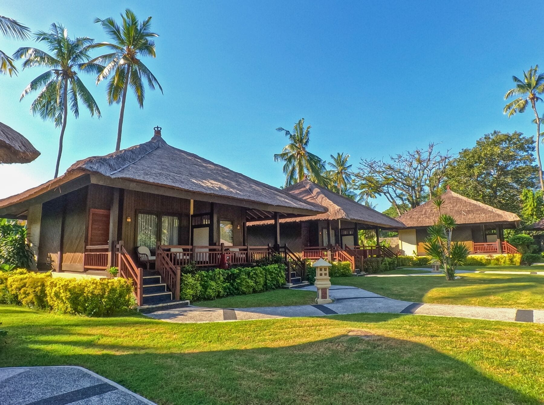 Un complexe hôtelier doté de bungalows aux toits de chaume, d’une pelouse bien entretenue et de palmiers en arrière-plan.