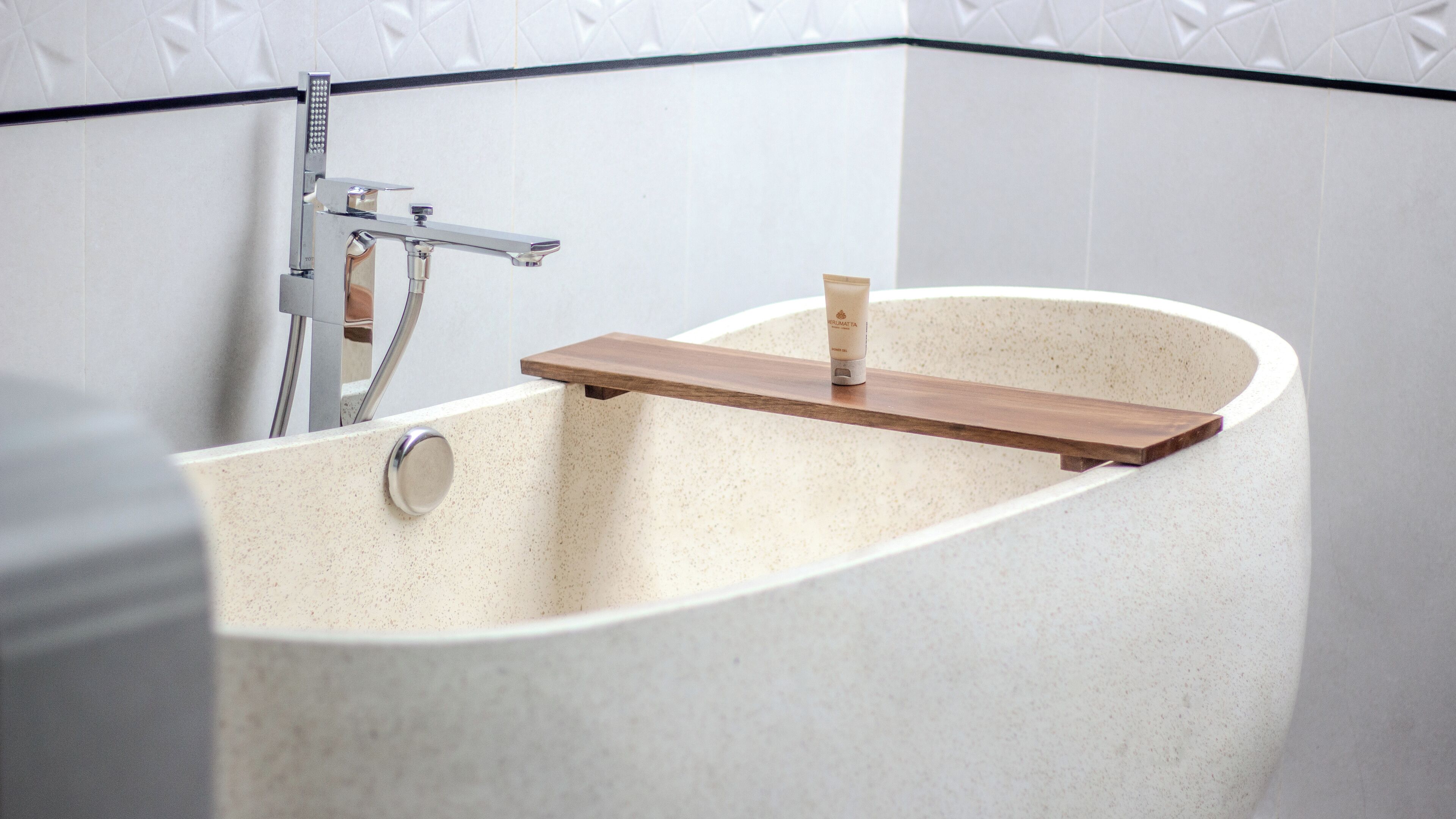 A bathtub with a wooden shelf, a chrome faucet, and a textured wall.