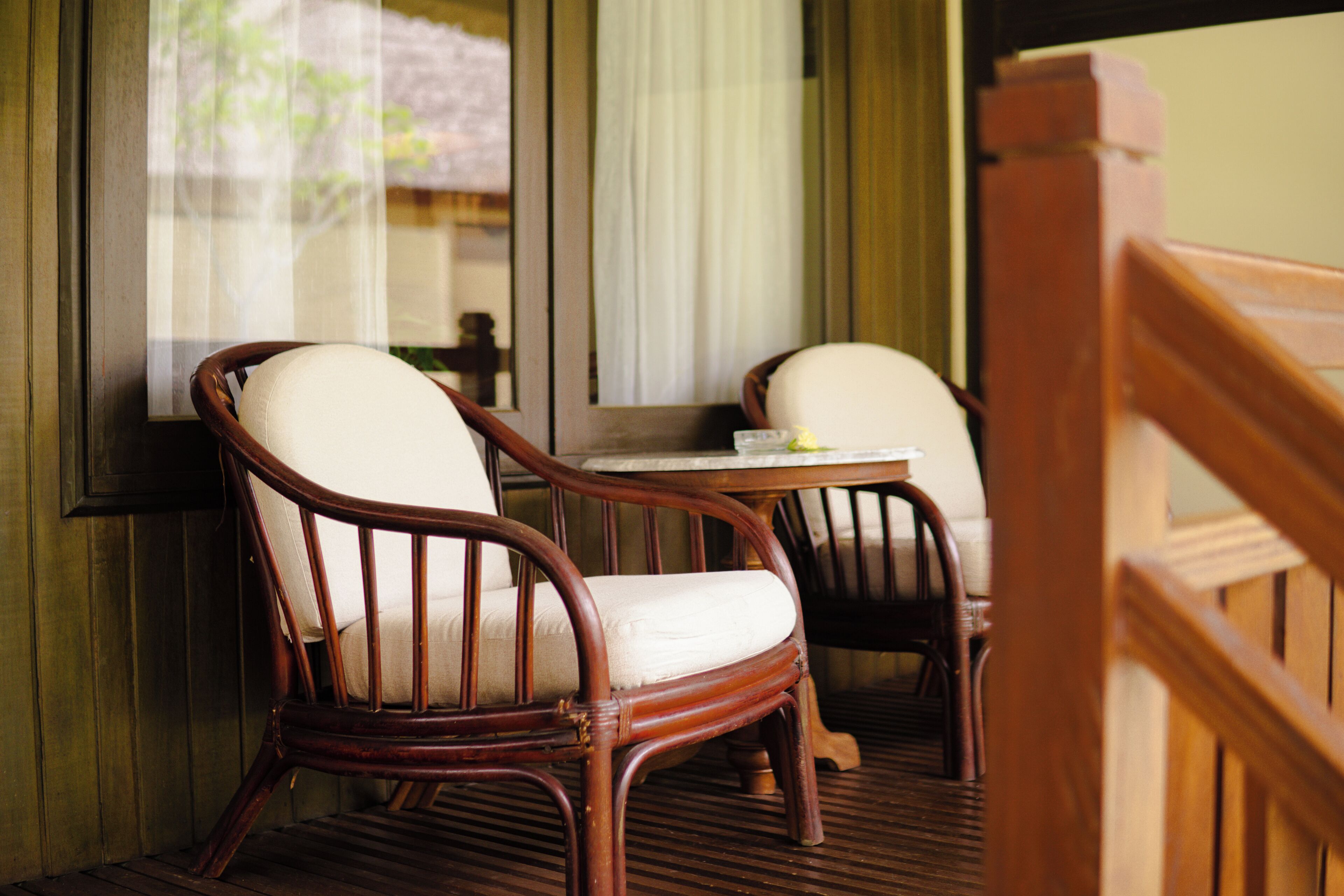 Two wicker chairs with white cushions and a small wooden table, positioned near a window with a view of greenery outside.