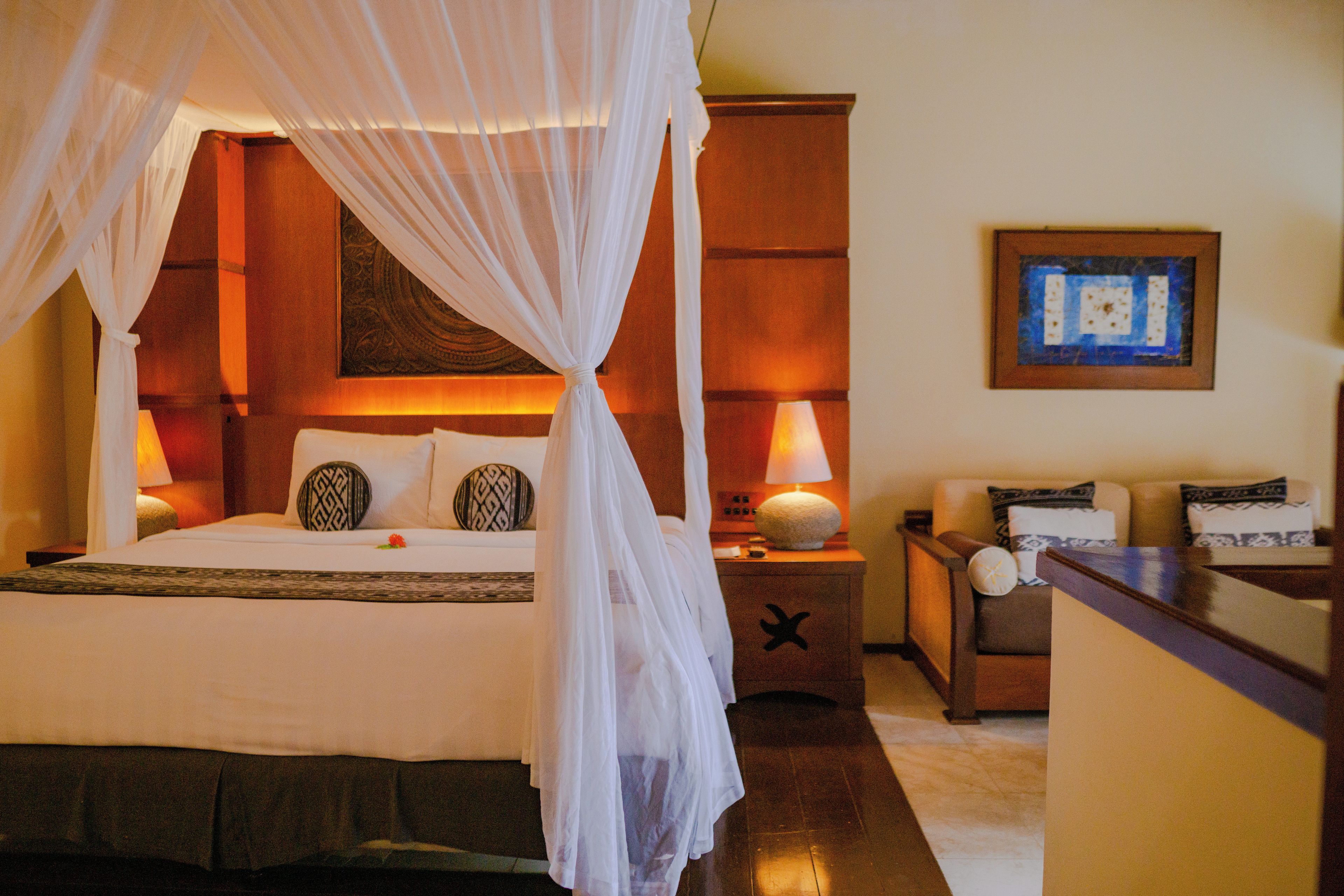 Habitación de hotel con cama con dosel, mesita de noche con lámpara y un cuadro en la pared.