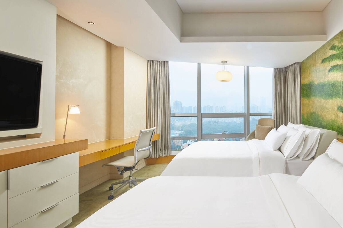 A hotel room with a large bed, a desk with a chair, a TV, and a city view through the window.