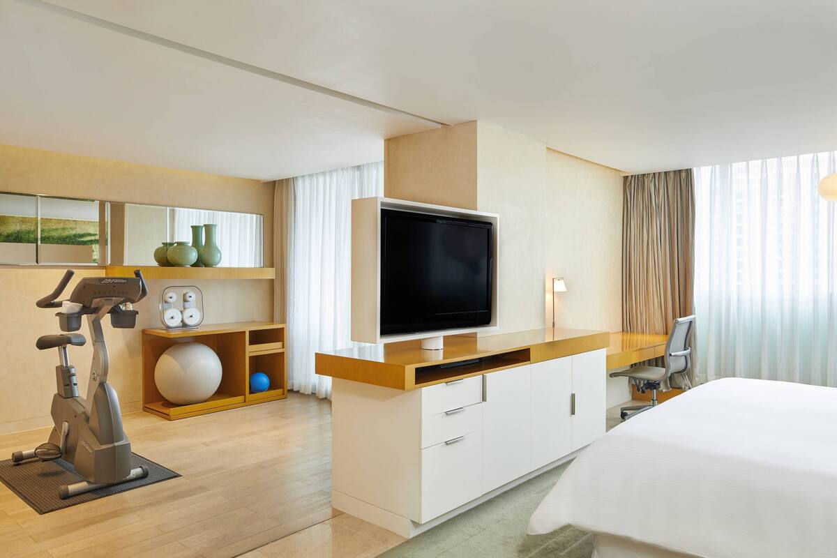 A hotel room with a bed, a TV, a desk with a chair, a treadmill, and exercise balls.