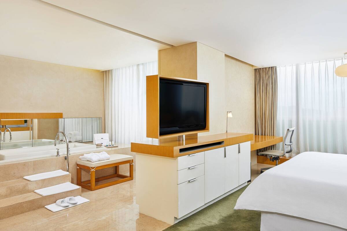 A hotel room with a large flat-screen TV, a bed, a desk with a chair, a bathroom with a sink and mirror, and a staircase.