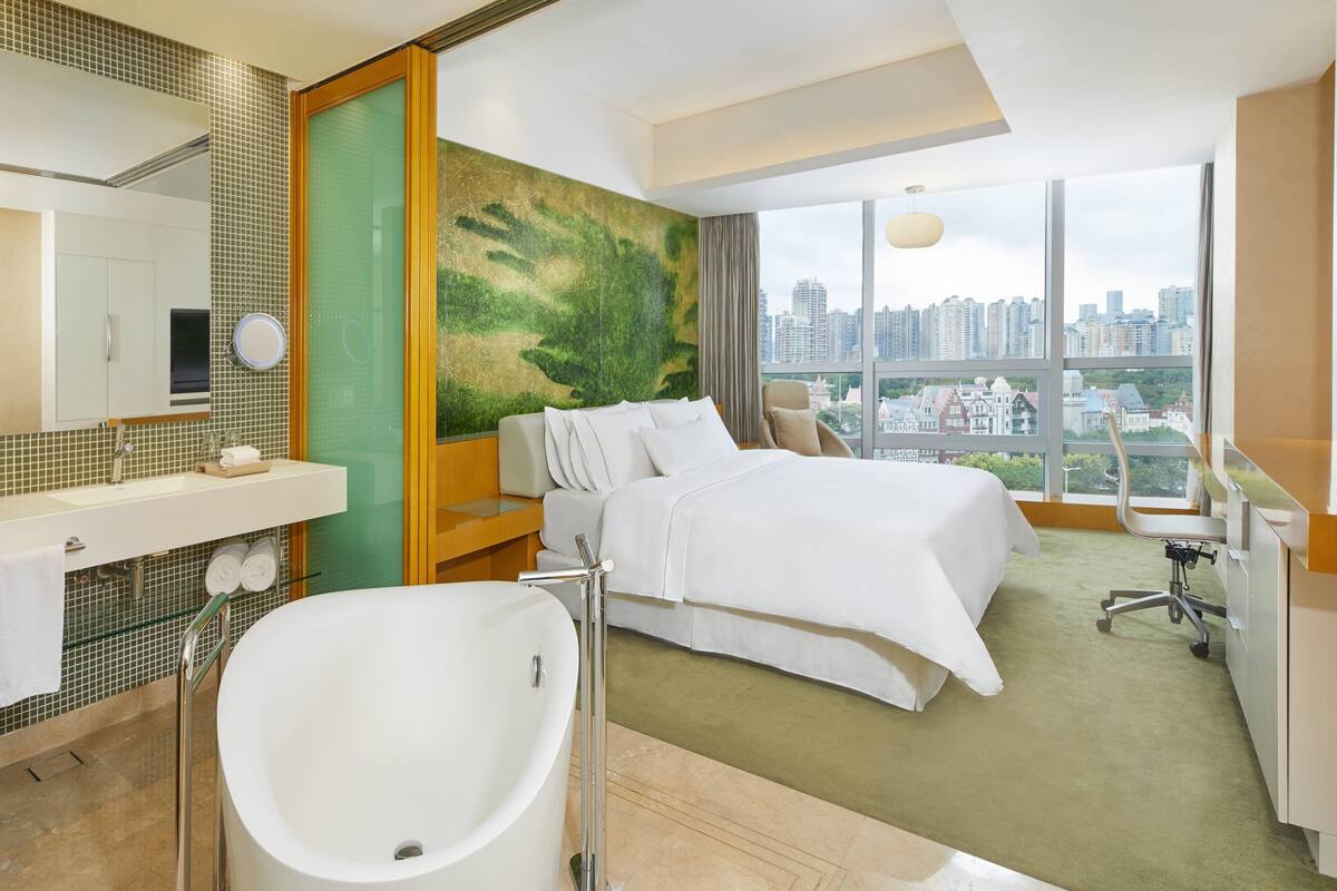 A hotel room with a large bed, a bathtub, a city view, and a modern bathroom.