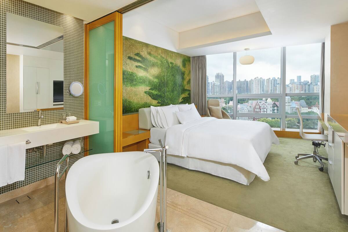 A hotel room with a large bed, a bathtub, a vanity with a mirror, and a city view through large windows.