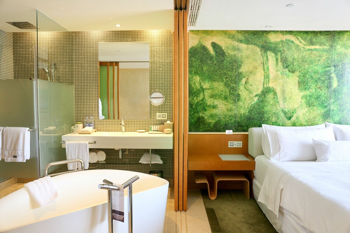A modern hotel room with a large bathtub, a separate shower area, a large mirror, a wooden vanity, a bed, and a green mural on the wall.