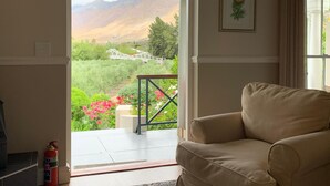 Ein Zimmer mit beiger Couch, einer Tür zu einem Balkon mit Blick auf Berge und Grünflächen, und einem gerahmten Pflanzenmotiv an der Wand.
