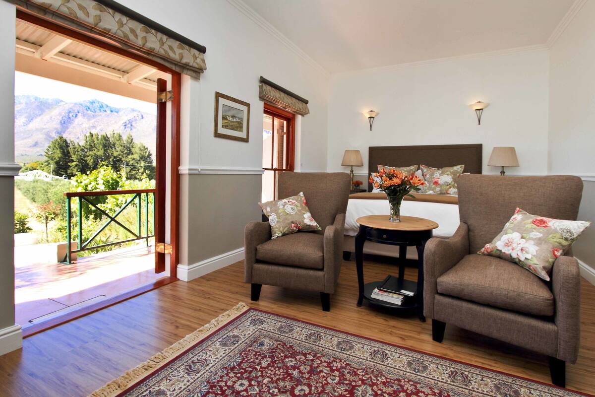 Een kamer met twee fauteuils, een bijzettafeltje en een bed met hoofdeinde. Er is een raam met uitzicht op bergen en een balkon met een hekwerk.