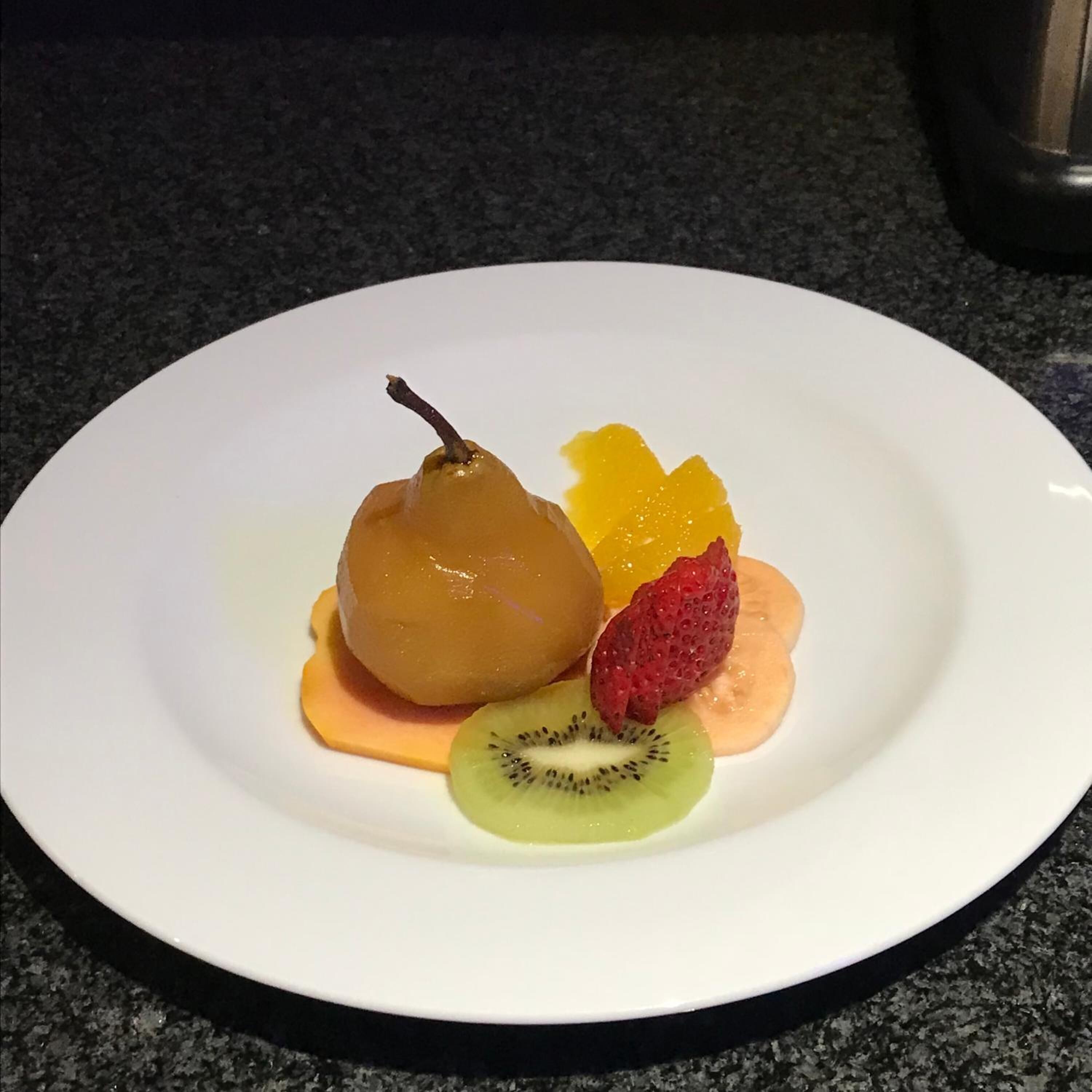 Ein weißer Teller mit einer Birne, einem Kiwistück, einer Erdbeere und einer Orangenscheibe auf einer dunklen Granitabdeckung.