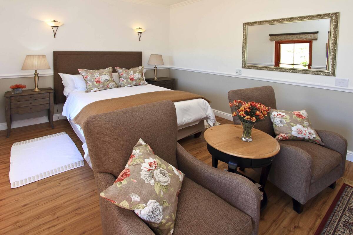 Een slaapkamer met een groot bed, twee nachtkastjes, een ronde houten tafel, twee fauteuils en een spiegel aan de muur.