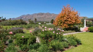 Ein Garten mit verschiedenen Blumen und einem Baum, ein Haus mit einer Veranda und Berge im Hintergrund.