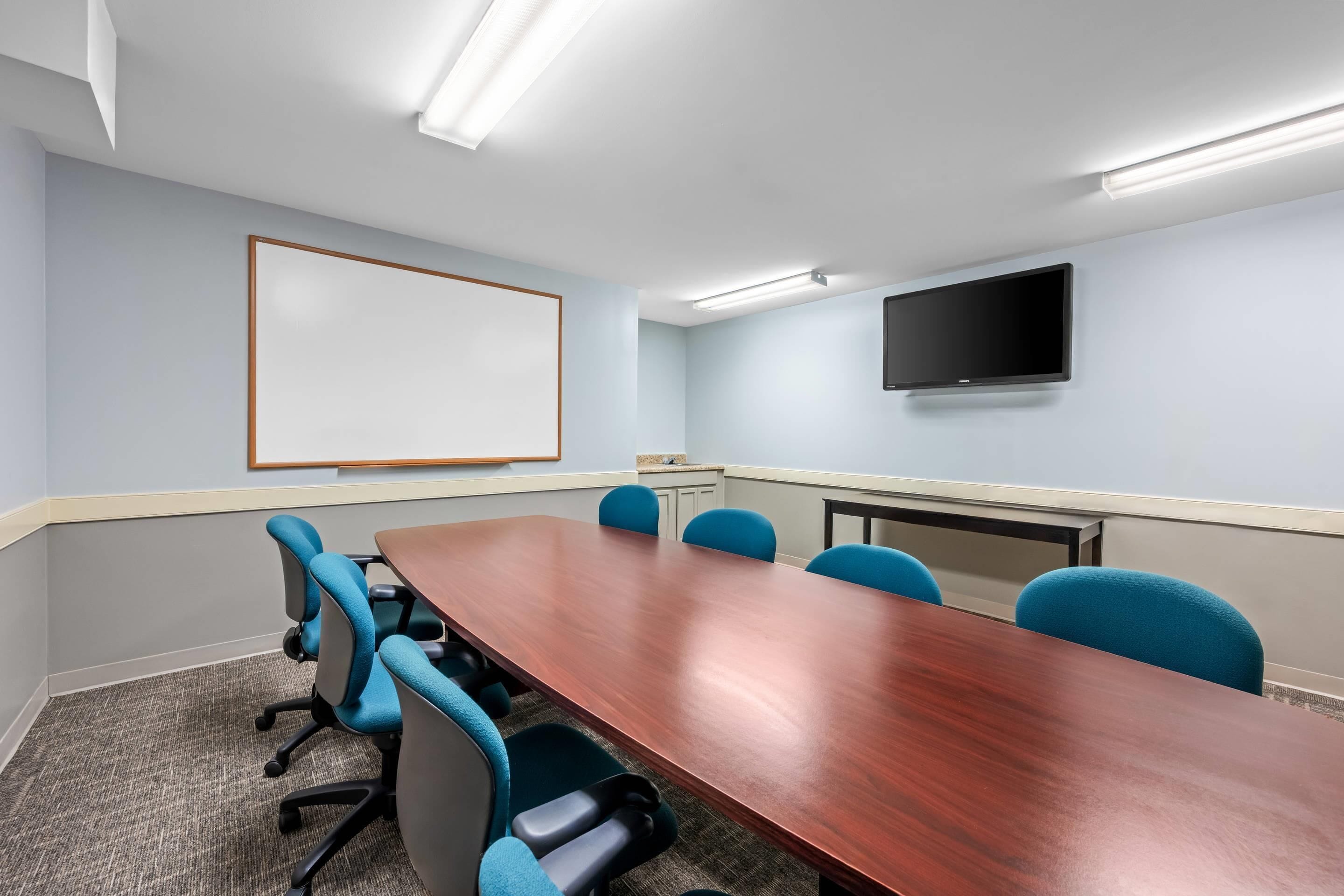 A conference room with a long wooden table, blue chairs, a whiteboard, and a flat-screen TV on the wall.