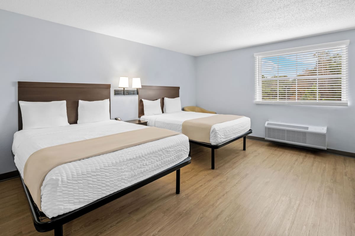 Une chambre d’hôtel avec deux lits, une fenêtre avec des stores, un radiateur fixé au mur et une table de chevet avec des lampes.