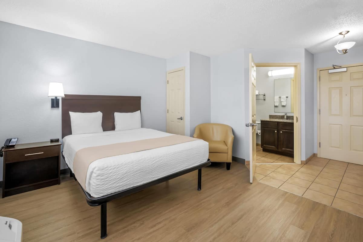 Une chambre d’hôtel avec un lit, une table de chevet, une chaise, une salle de bain visible à travers une porte ouverte et un plancher en bois.