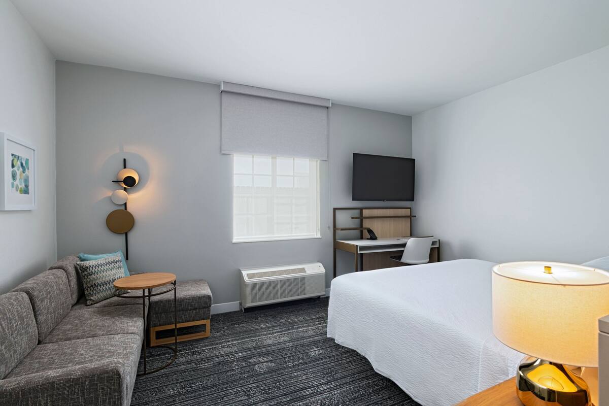 A hotel room with a bed, a desk, a sofa, a lamp, a TV, and a window with blinds.