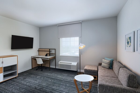 Una habitación compacta con escritorio, sofá, una mesita, una lámpara de pie y un televisor montado en la pared.