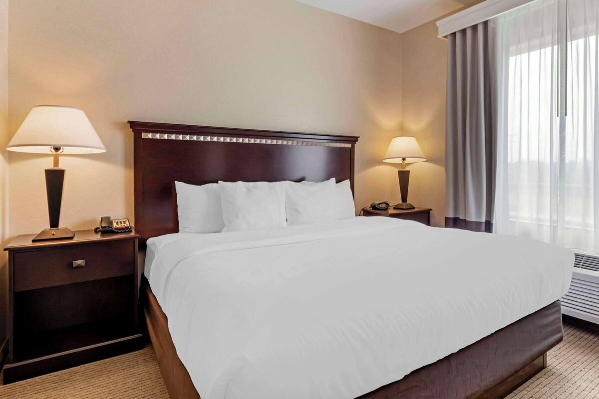 A hotel room with a large bed, two bedside tables with lamps, a nightstand, and a window with curtains.