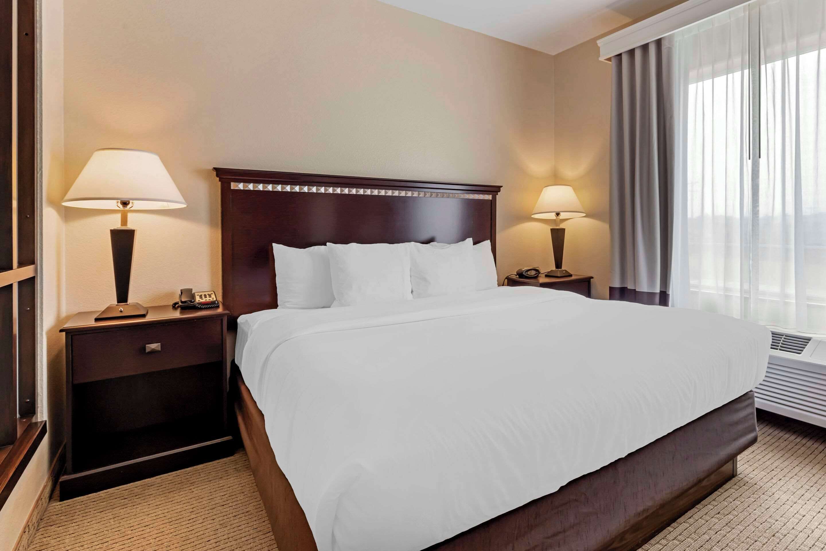 A hotel room with a large bed, two bedside tables with lamps, a nightstand, and a window with curtains.