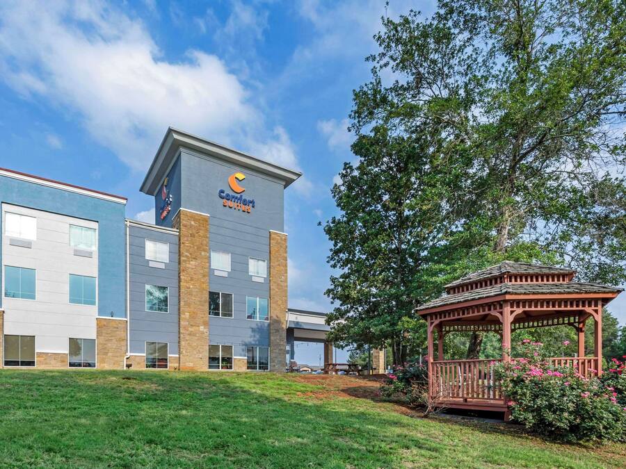 A Comfort Suites hotel with a modern design, featuring a large glass facade and a well-maintained garden with a gazebo.