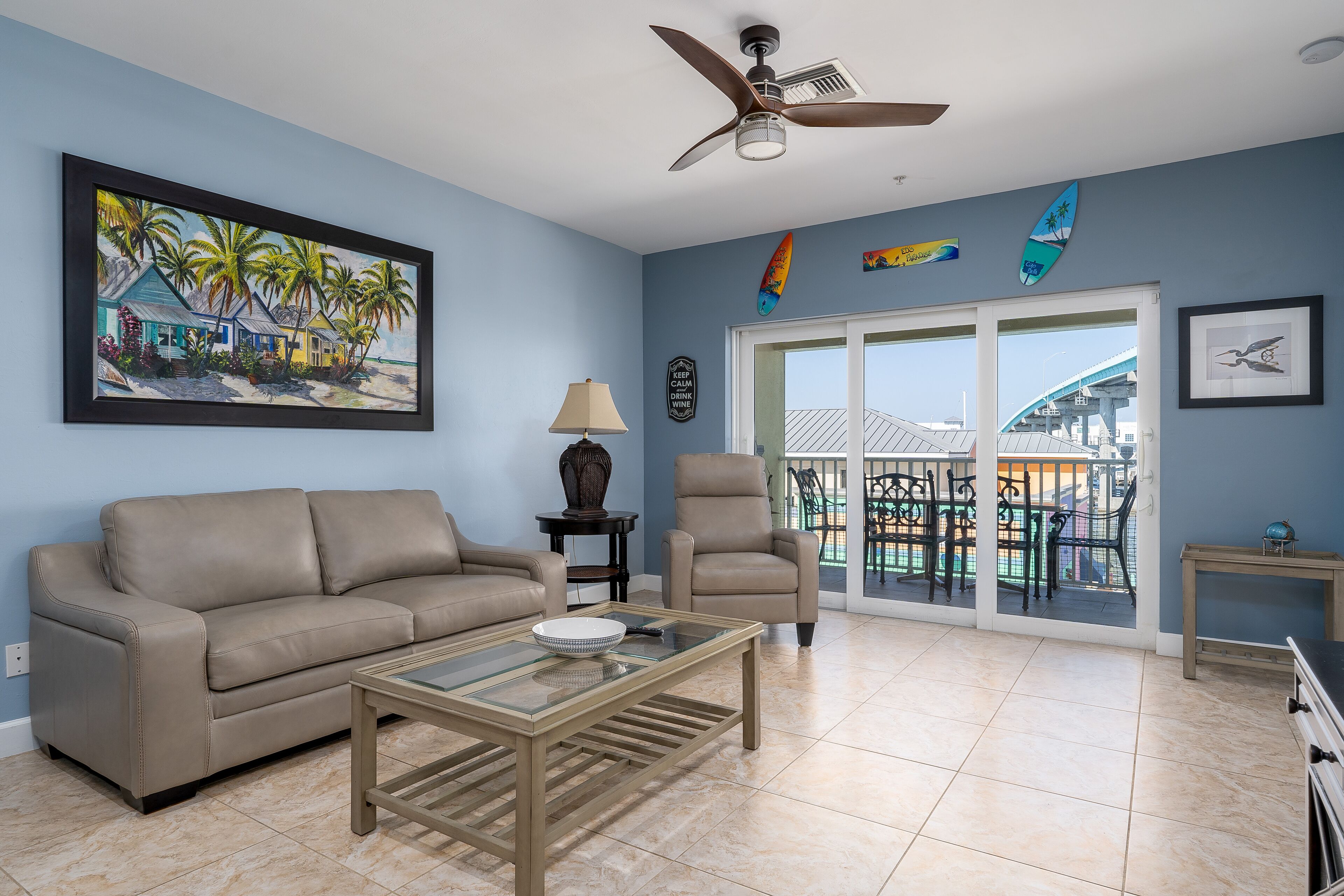 A living room with a beige sofa, a glass coffee table, a ceiling fan, and a large painting of a beach scene.