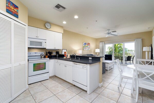 A modern kitchen with white cabinets, a microwave, an oven, and a dishwasher. There is a dining area with a table and chairs, and a view of palm trees through the window.