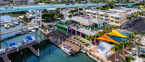 A marina with boats docked, a bridge overhead, and buildings with outdoor seating.