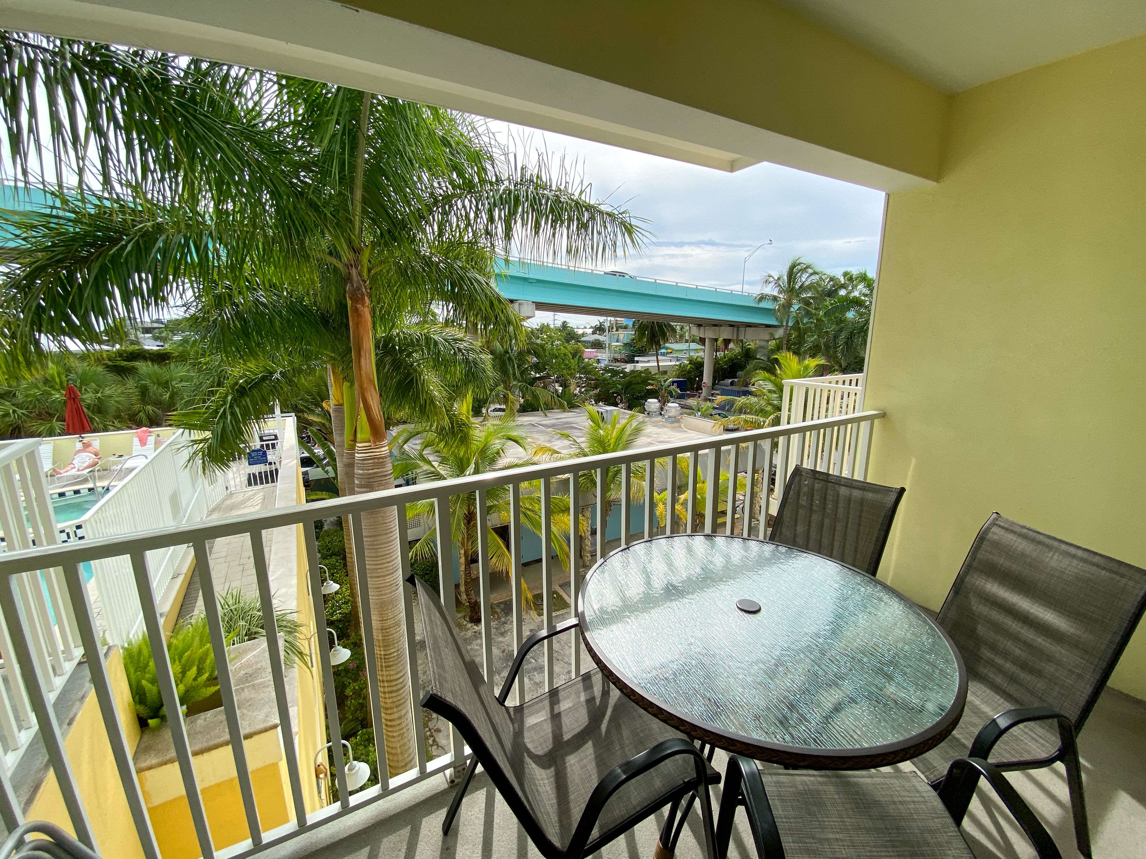 Un balcone con tavolo e sedie in vetro, con vista su una piscina e palme.