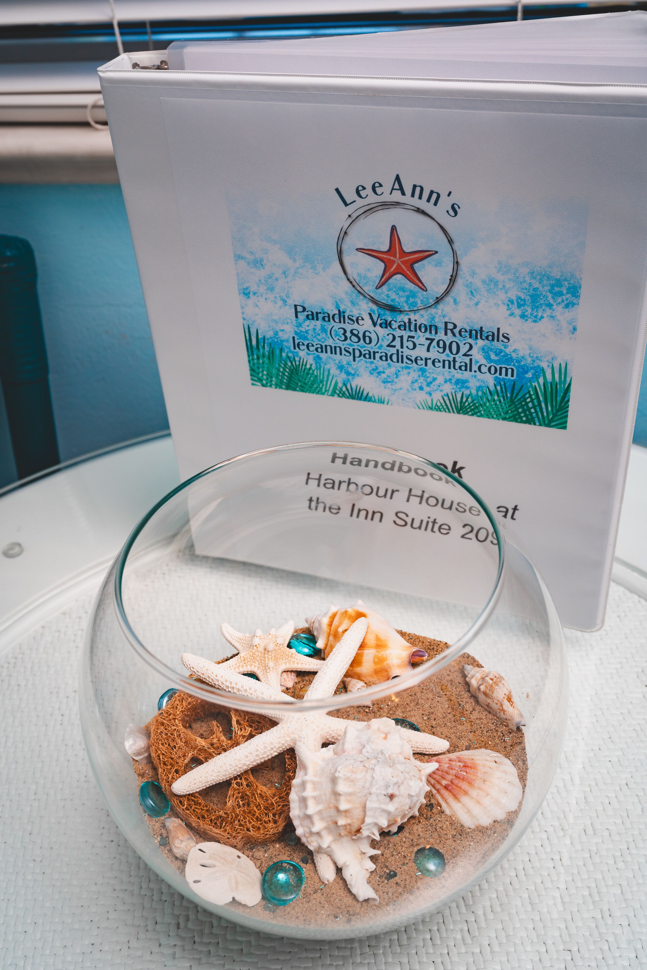 Un cuenco de vidrio con caracoles y estrellas de mar sobre una mesa, con un folleto de LeeAnn's Paradise Vacation Rentals al fondo.