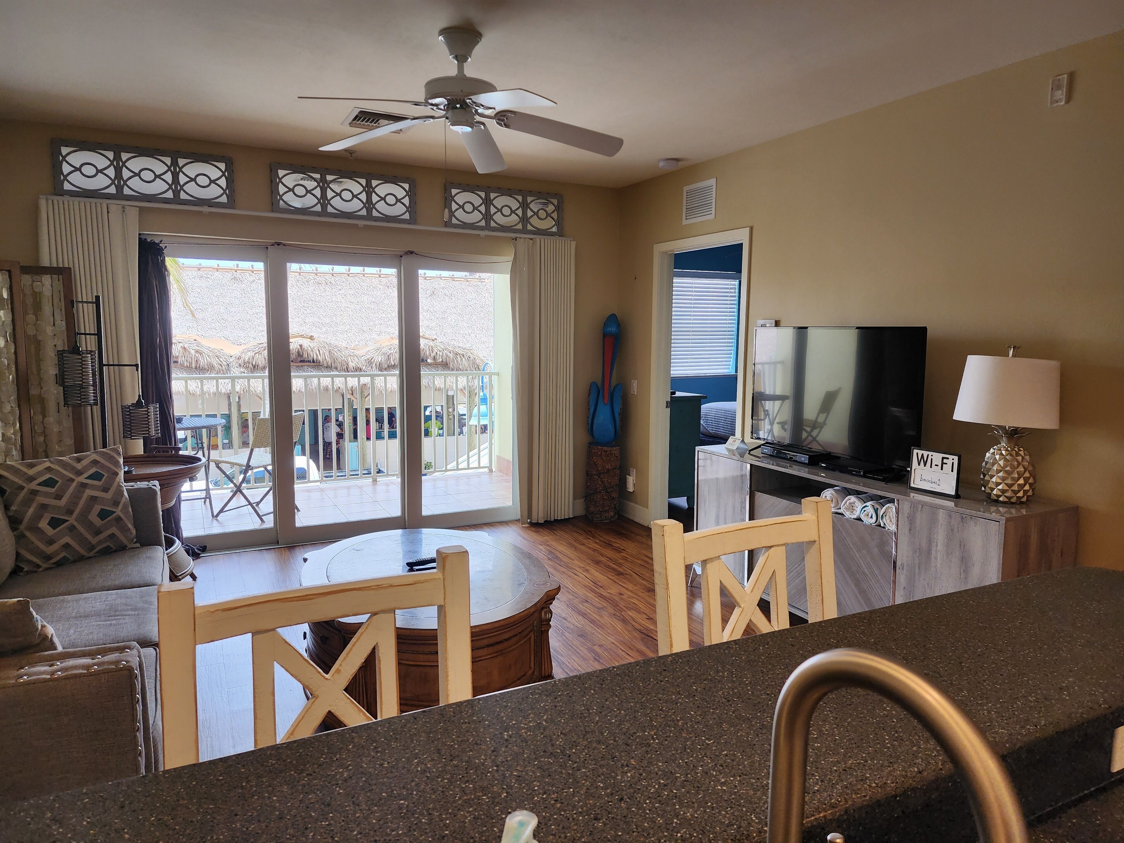 Un salon équipé d’un téléviseur, d’un ventilateur de plafond, d’une table à manger et offrant une vue sur le balcon extérieur.